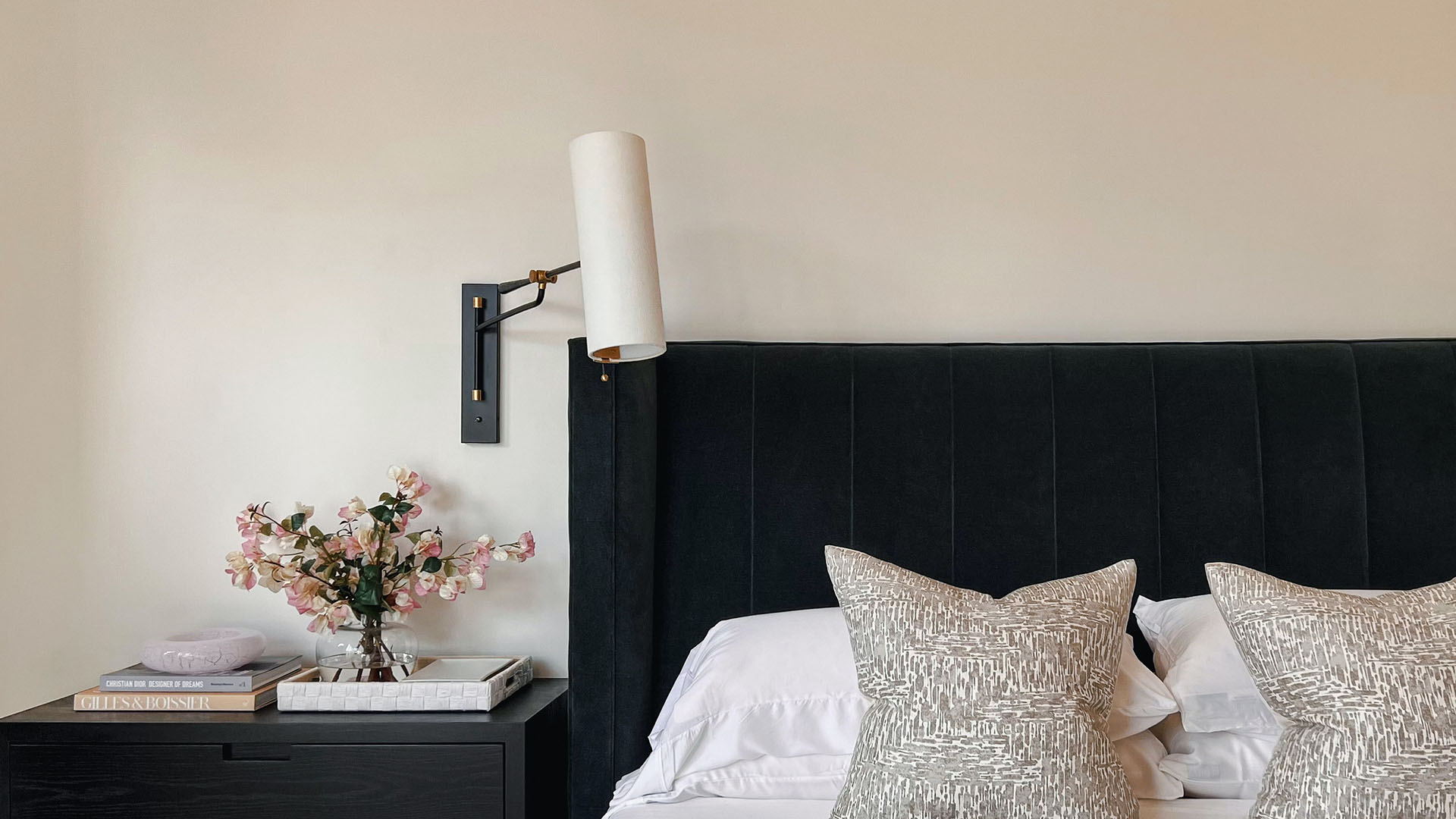 Bedroom with plush bed with headboard, bedside dresser with books and plants, and lamp.