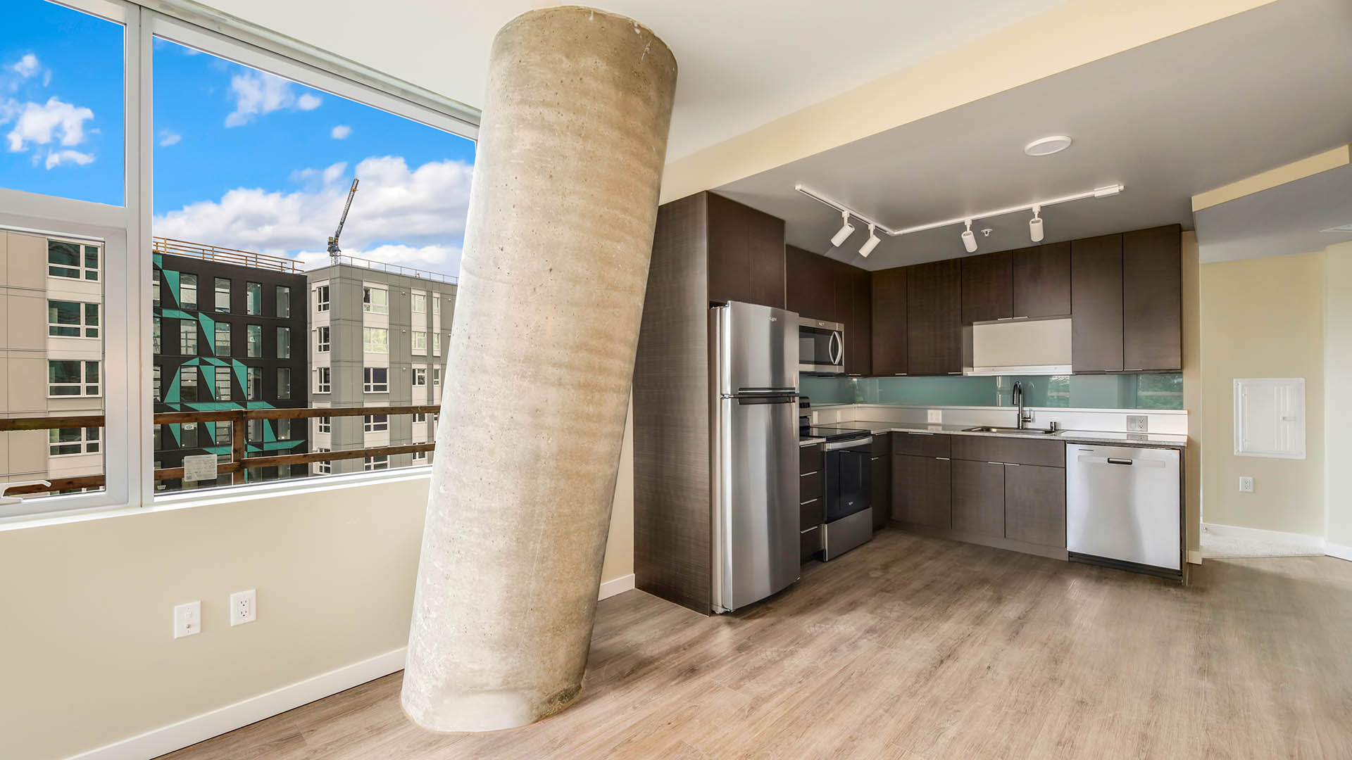 Apartment kitchen with wood style floor and cabinets, stone counters, stainless steel appliances, and large concrete pillar.