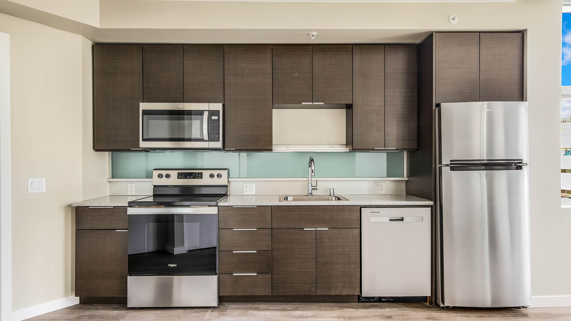 Apartment kitchen with wood style floor and cabinets, stainless steel appliances, and blue accent backsplash.